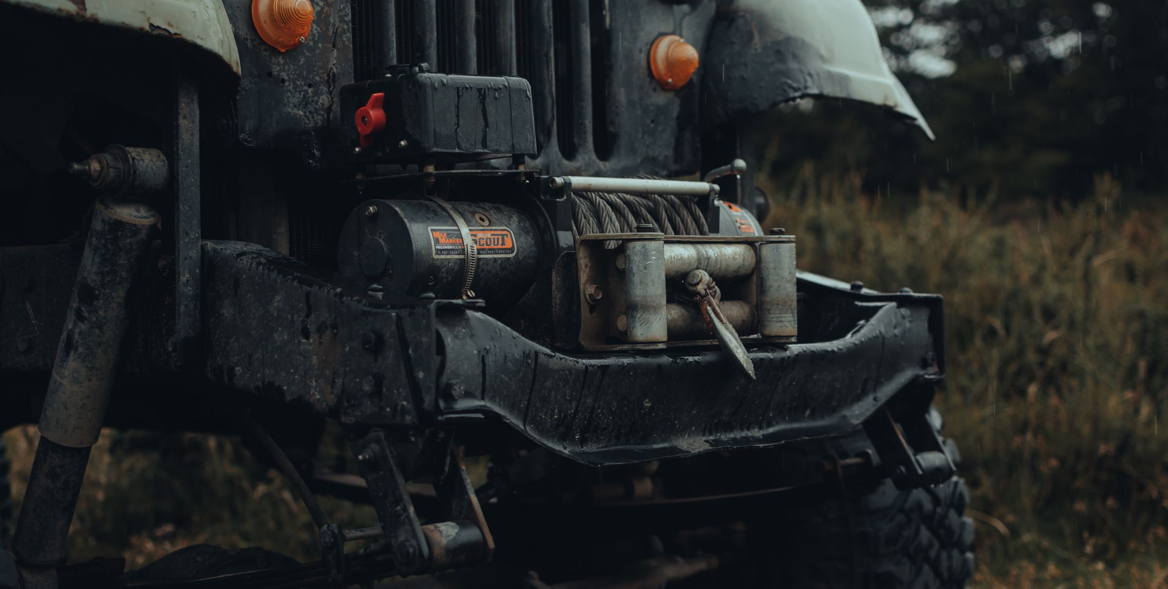 Close-up of a front-bumper-mounted winch with steel cable on a black off-road truck in wet conditions