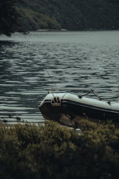 A tranquil scene of a fishing boat docked by a forested lake under light rain, capturing peaceful outdoor leisure.