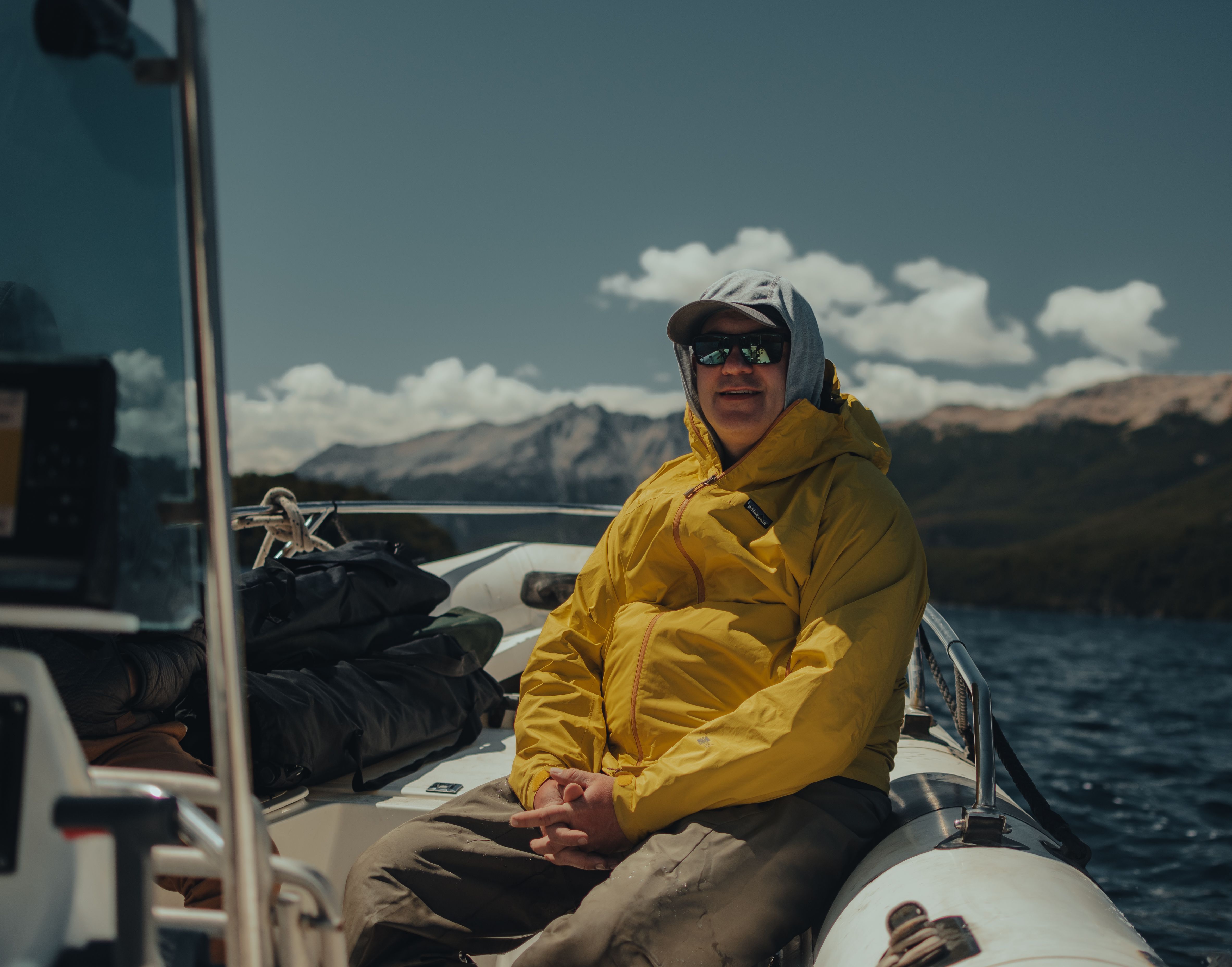Adult male in yellow jacket on a boat, embracing a sunny day at sea with mountains in the background.