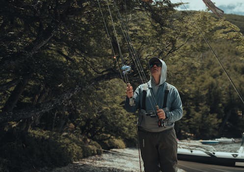 A person prepares to fish by a tranquil, forested lakeside during the day.