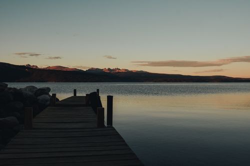 Free Tranquil evening at a calm lake with a wooden dock and mountain silhouette during sunset. Stock Photo
