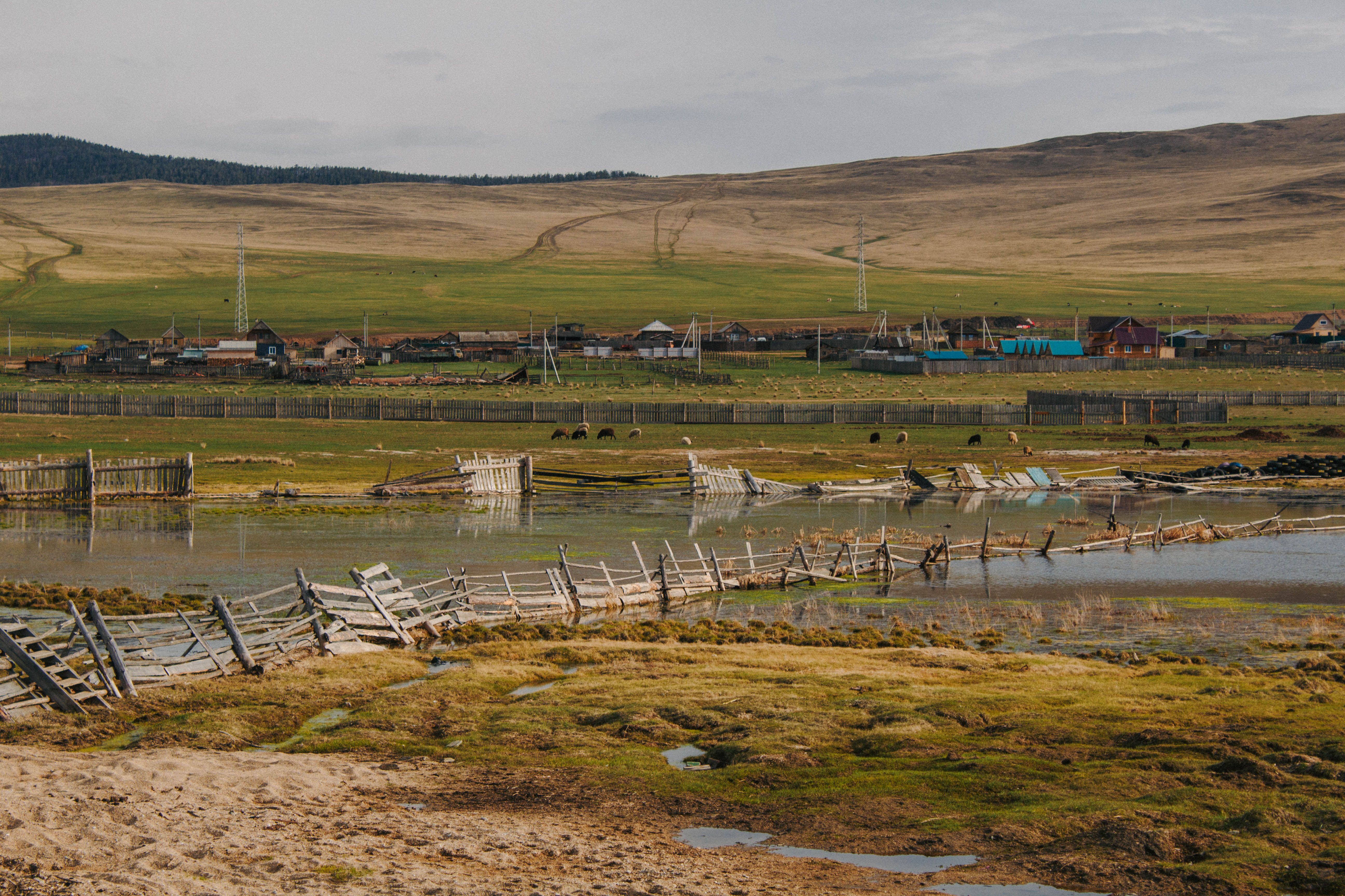 Scenic view of a rural landscape featuring a dilapidated wooden fence and a tranquil pond near a distant village.