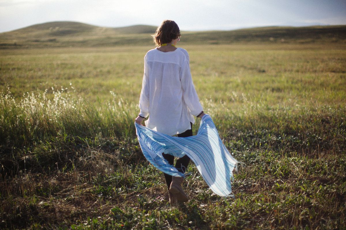 A woman seen from behind, crossing a sunlit grass field with a scarf trailing