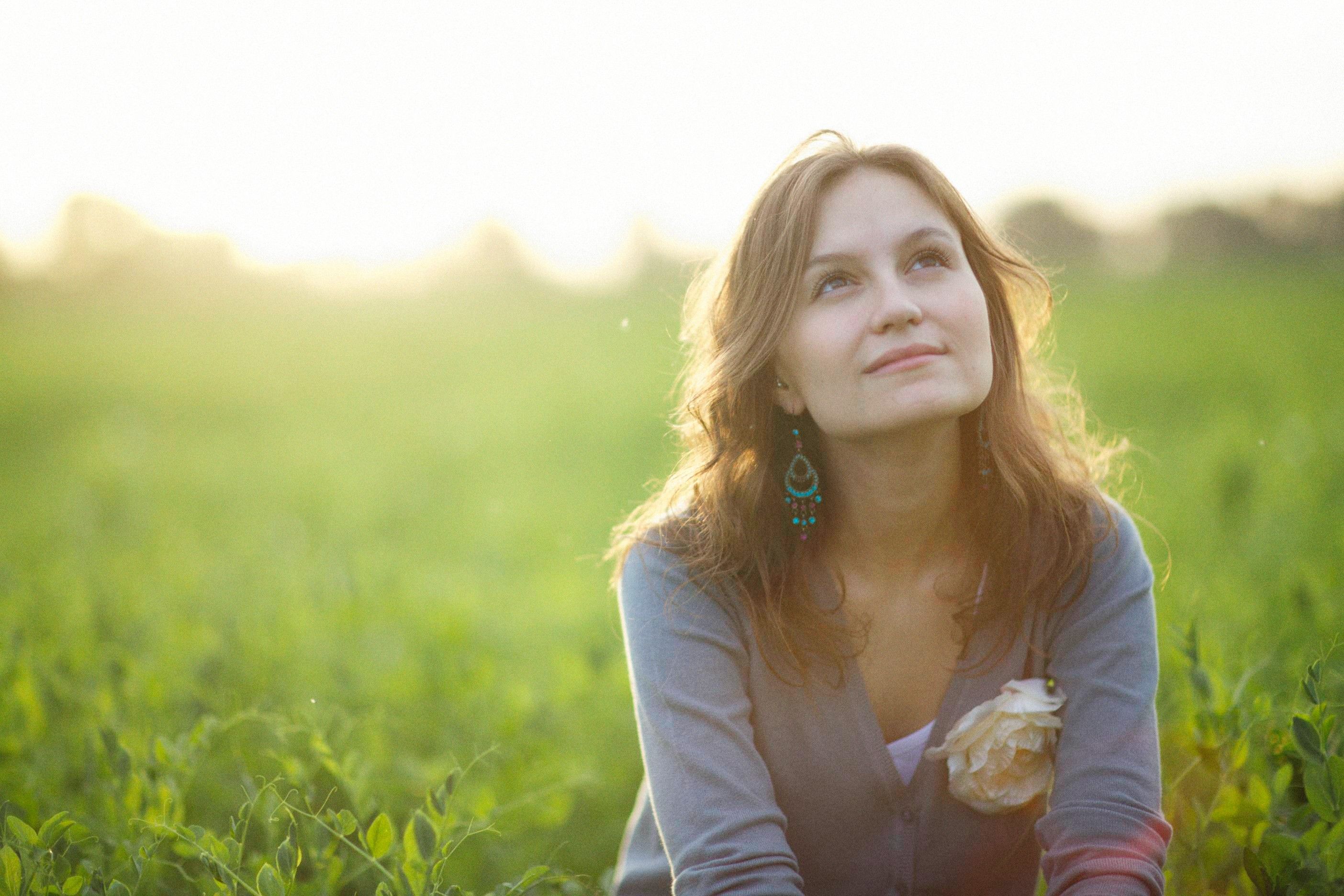 A carefree woman sitting in a sunlit field looks upward with a peaceful smile.
