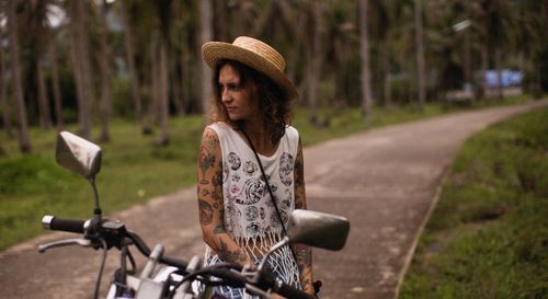 Free Tattooed woman in a straw hat standing by a motorcycle on a rural path. Stock Photo