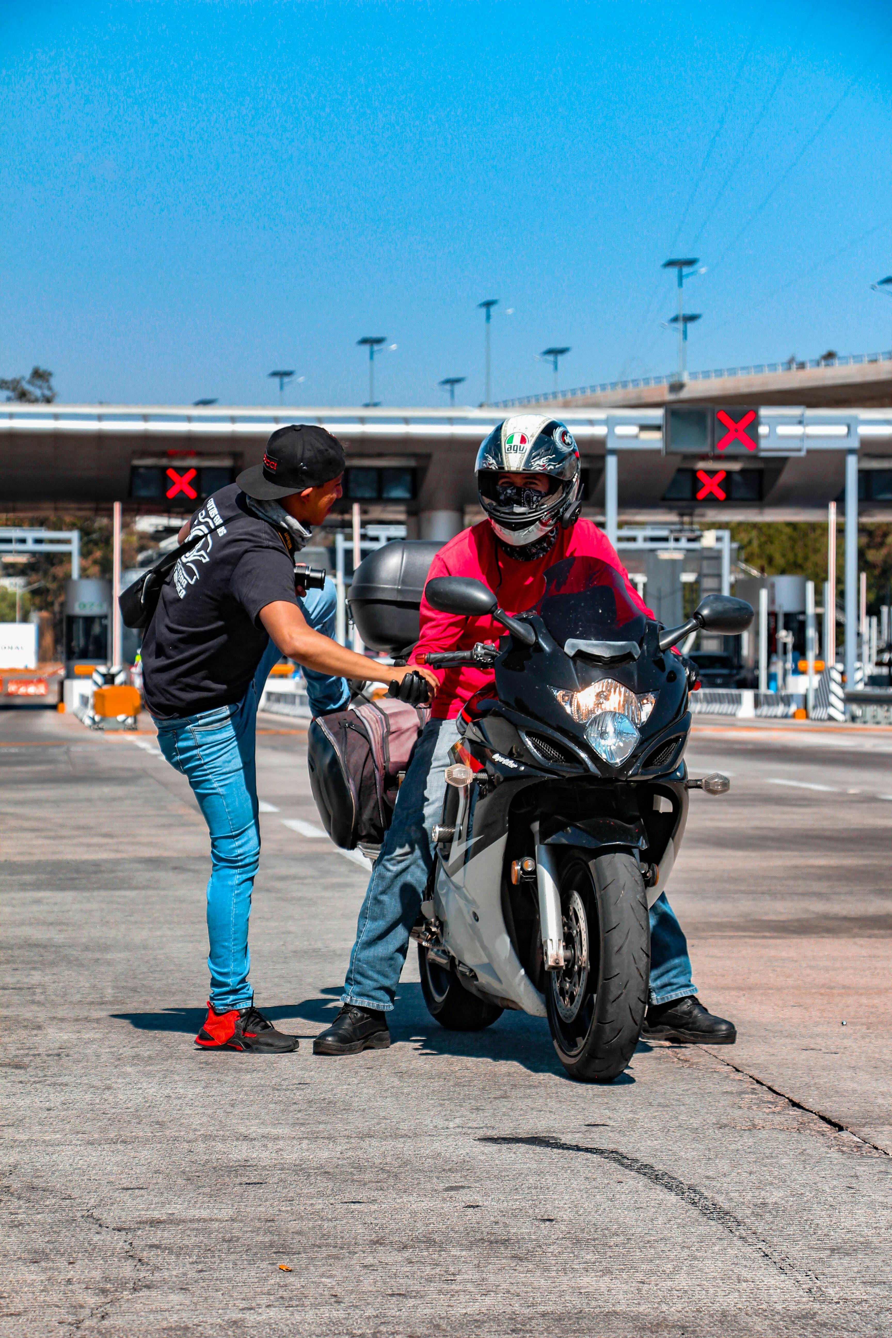 Two men at a toll booth, a motorcyclist receiving a bag from a friend.