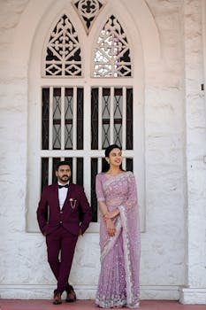 Stylish couple posing outdoors in traditional attire beside a Gothic window.