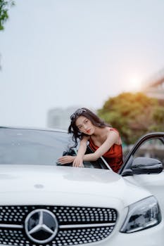 Asian woman in red top leaning on a white Mercedes car, exuding elegance and style.