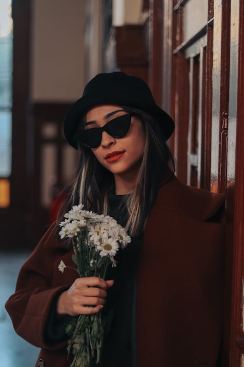 Close-Up Shot of a Woman in Brown Coat and Black Fedora Hat Wearing Sunglasses while Holding White Flowers