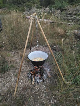 Outdoor cooking pot suspended over a campfire in a rustic setting with green foliage