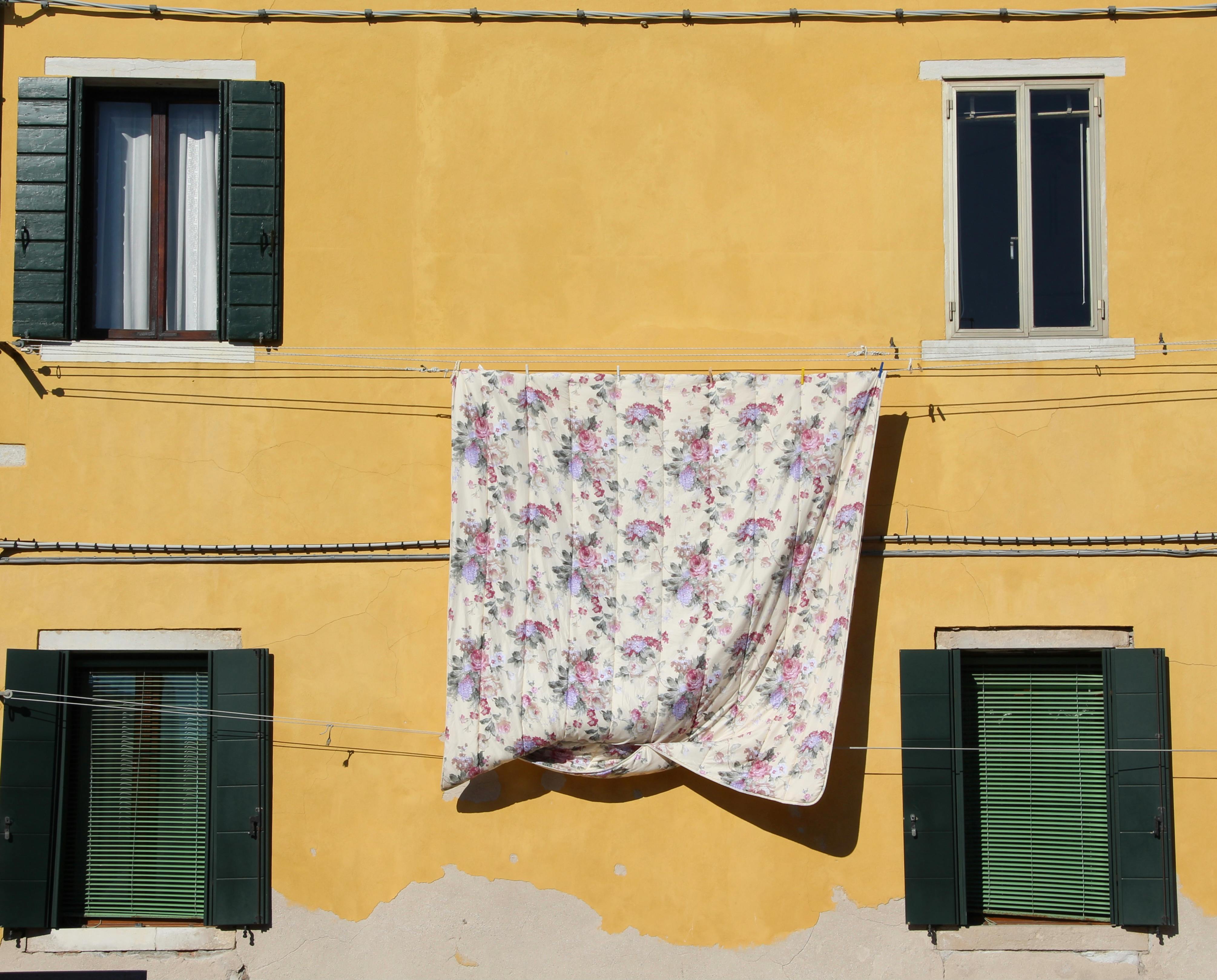 A floral blanket dries on a vibrant yellow building facade with green shutters and windows.