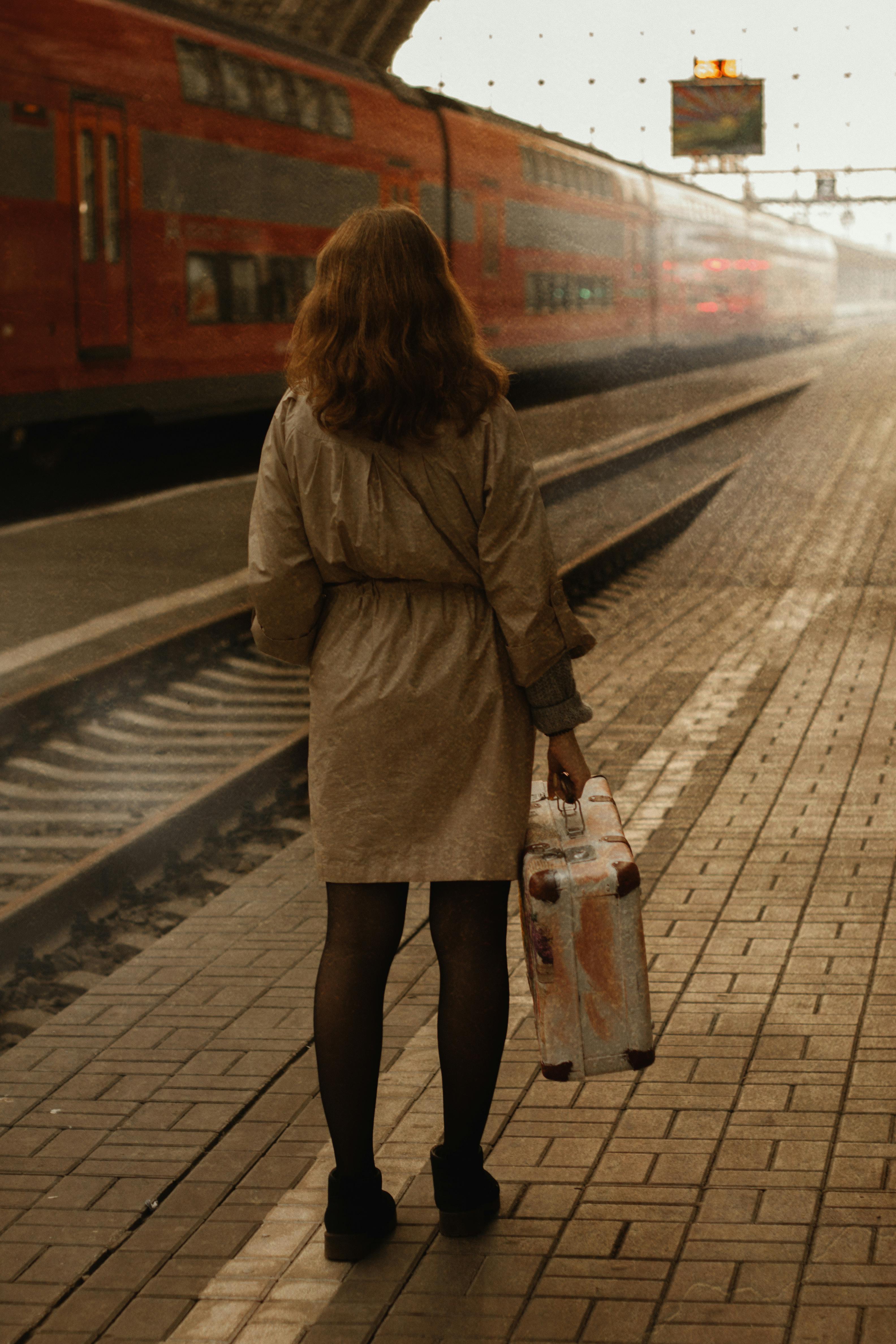 Woman standing with suitcase on railway platform as train arrives, creating a nostalgic travel scene.