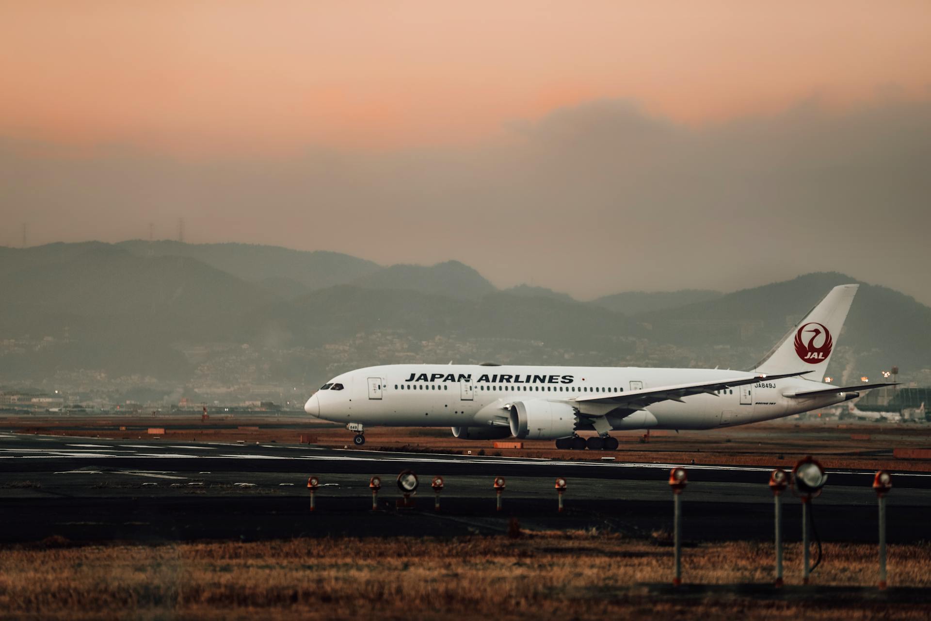 A Japan Airlines aircraft is taxiing on the runway during a scenic evening with mountains in the background.