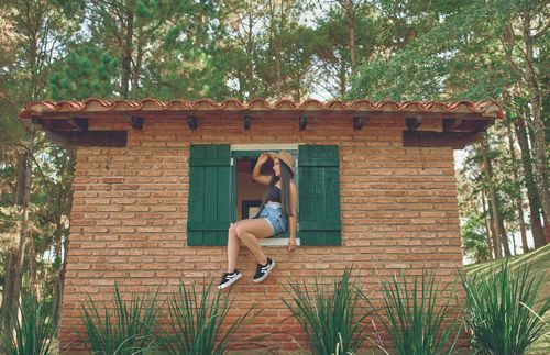 Free Woman in hat sitting in a window of a brick house, surrounded by nature. Stock Photo