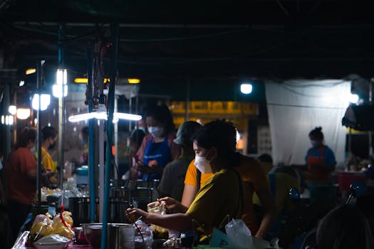 Vendors at a bustling night market wearing face masks, illuminated by bright lights.