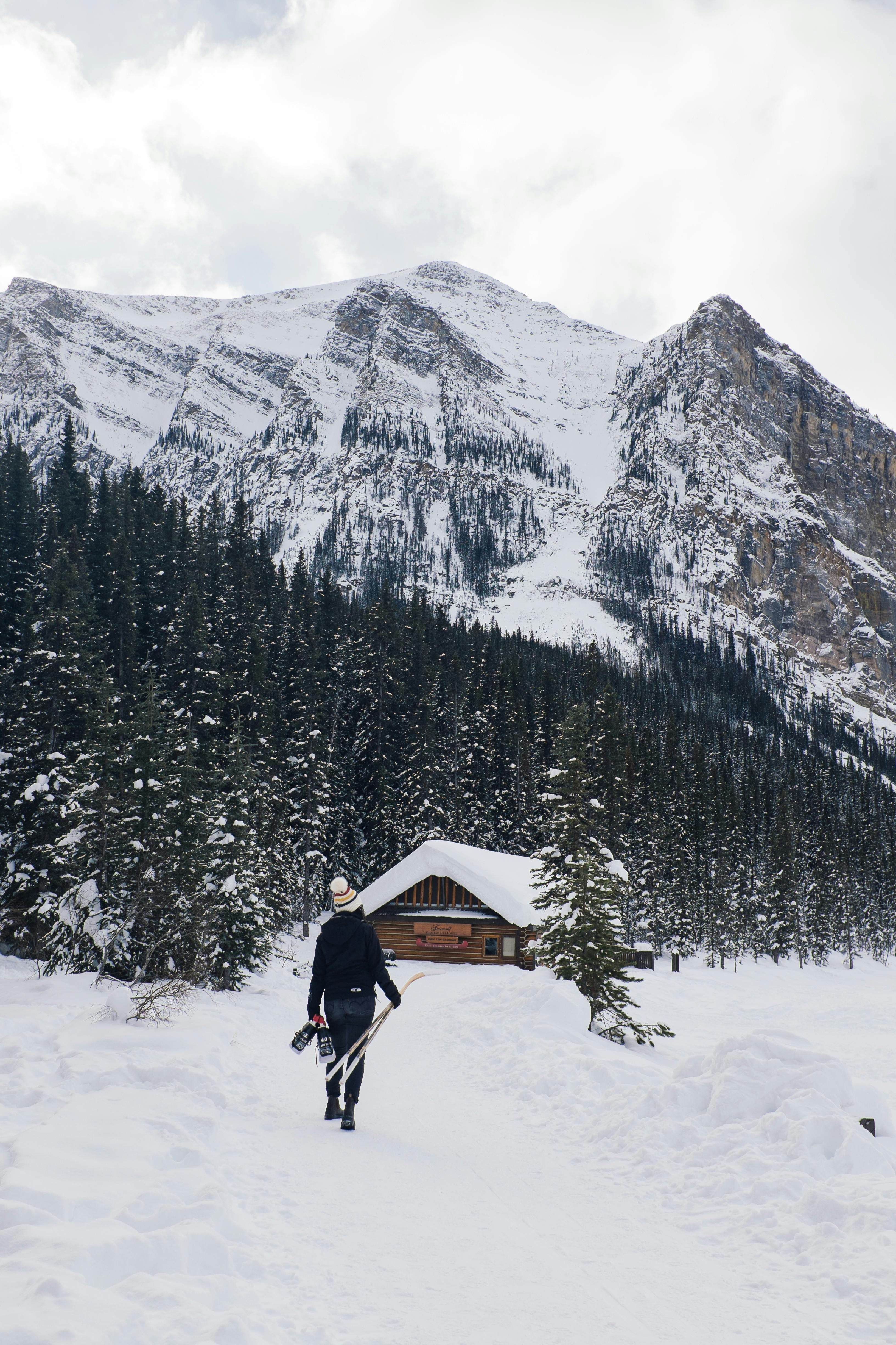 A person walks towards a cabin with snowy mountains and trees at Lake Louise, Canada.