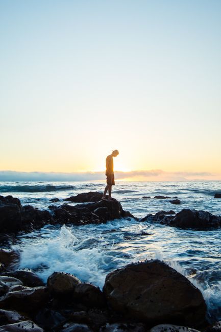 A tranquil scene of a man standing on rocks by the sea during sunset in Cape Town, South Africa.