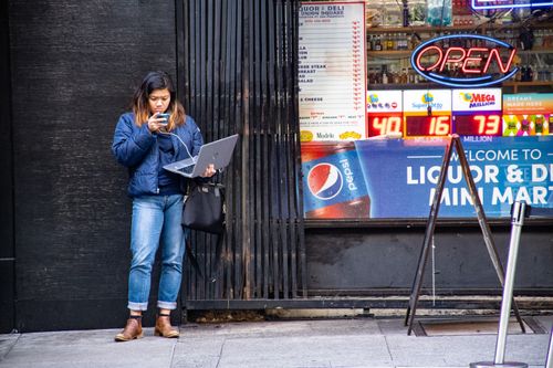Free Asian woman using a laptop and phone outside a city store by an open sign. Stock Photo