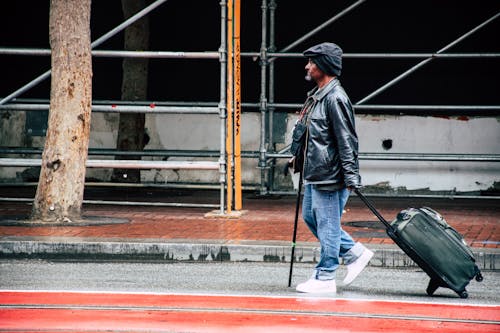 Free Walking Man in Black Leather Jacket and Blue Denim Pants Holding Luggage Bag Stock Photo