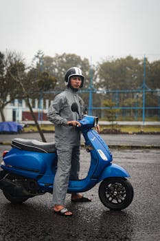 Man in raincoat stands beside a blue Vespa scooter on a rainy day outdoors.