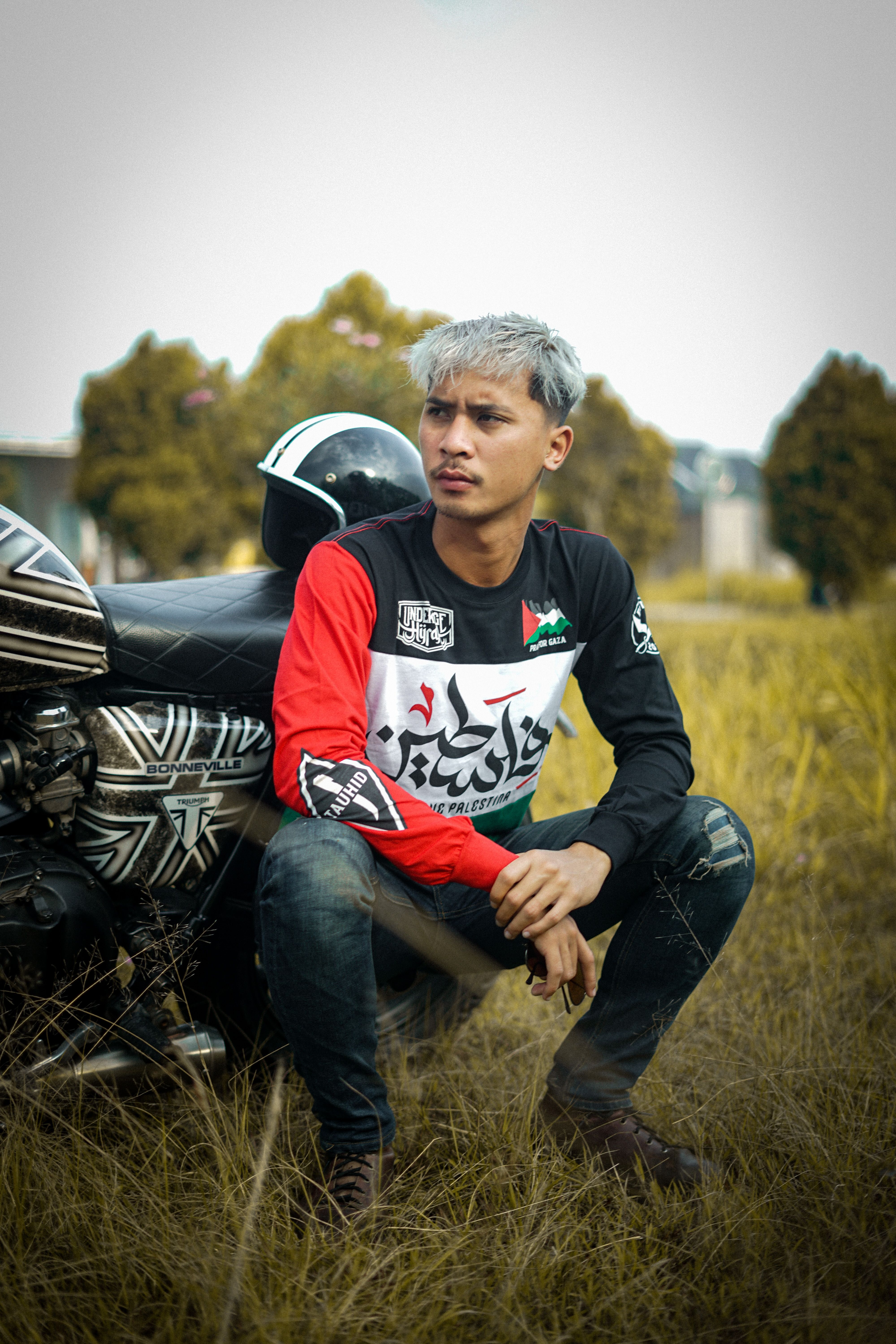 Stylish young man with dyed hair sits by a motorcycle in a grassy field.