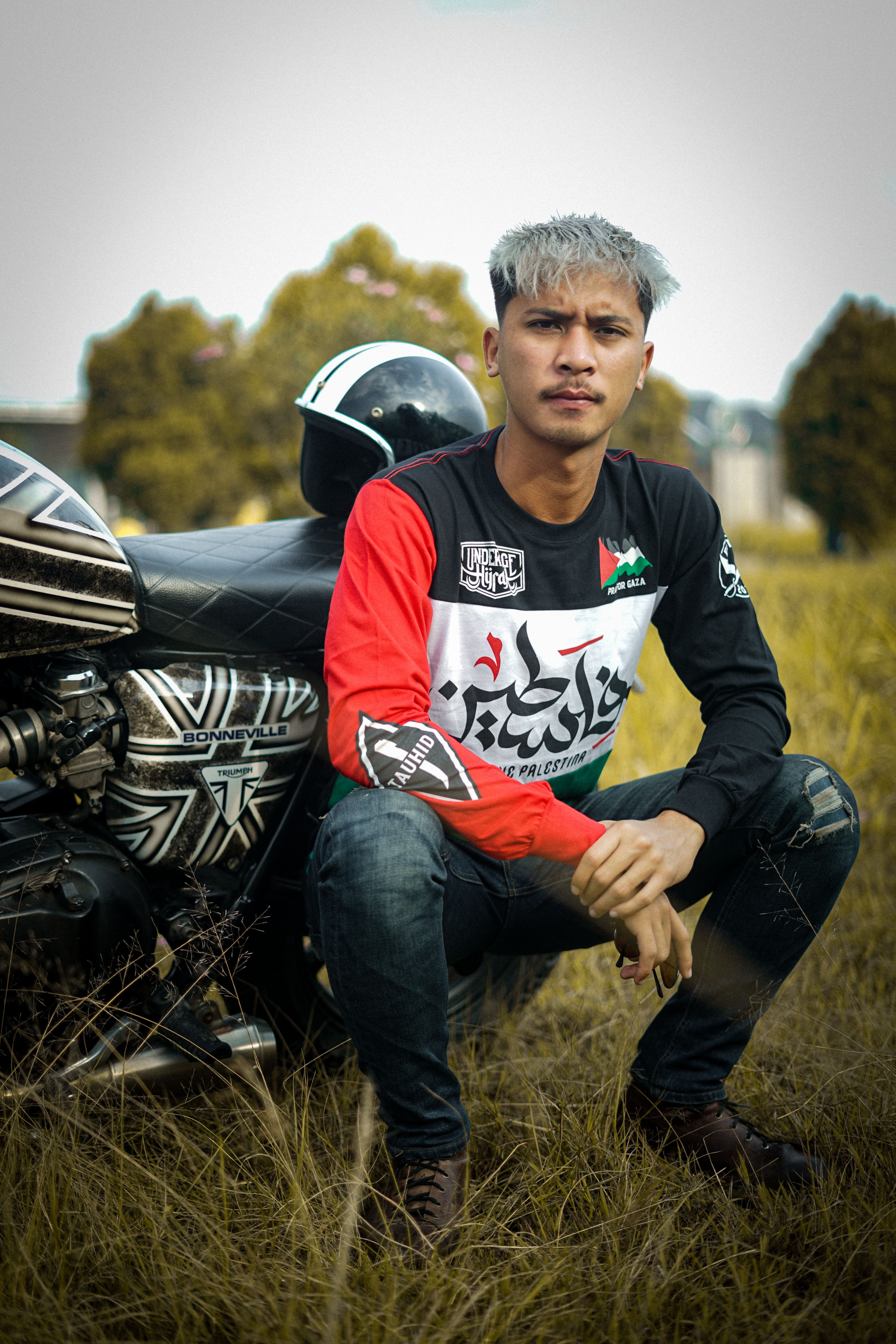 Confident young man seated beside a motorcycle in a grassy field, showcasing a stylish look.
