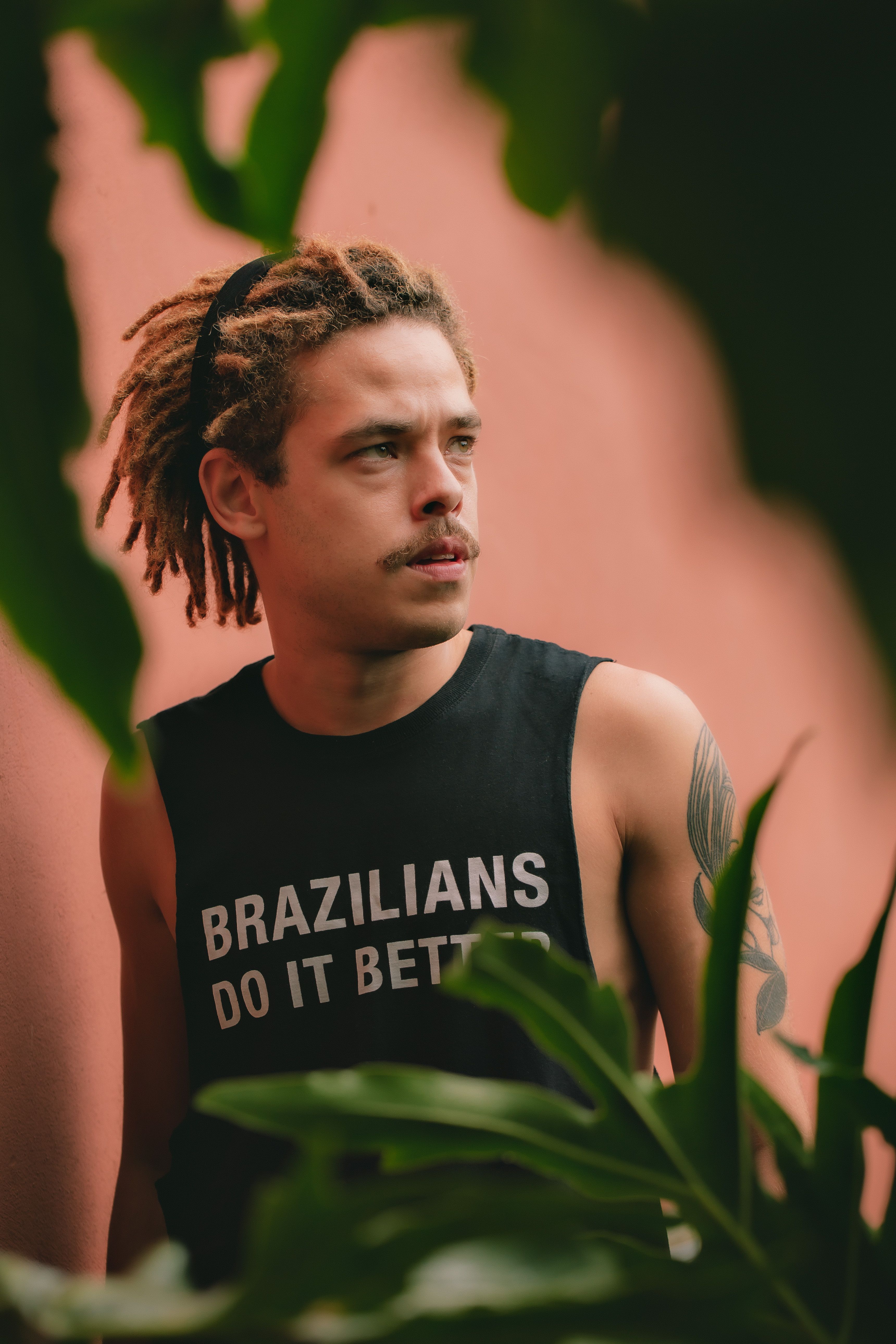 Confident man with dreadlocks and tattoo in sleeveless shirt stands outdoors.