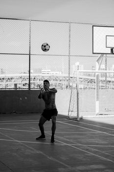 Black and white photo of a man playing soccer on a rooftop court with cityscape in the background.