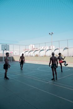 Four young men playing soccer on an outdoor basketball court in a city setting under clear blue skies.