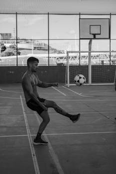 Shirtless man practicing soccer training on outdoor basketball court in black and white image.