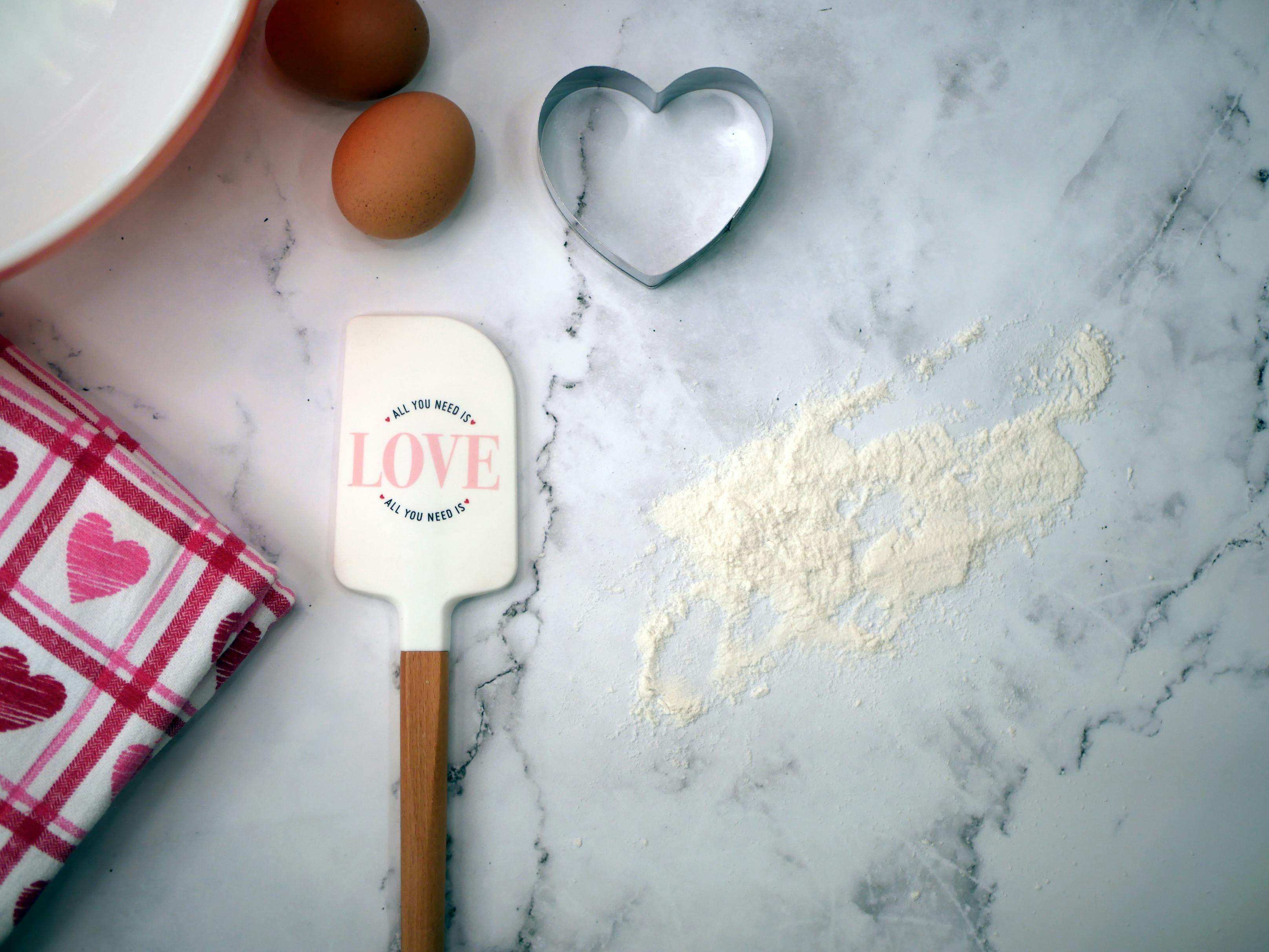 Heart-themed baking scene with eggs, heart cookie cutter, and 'Love' spatula on a marble surface.