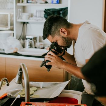 A male photographer takes close-up shots of food in a kitchen setting, focusing on presentation and detail.