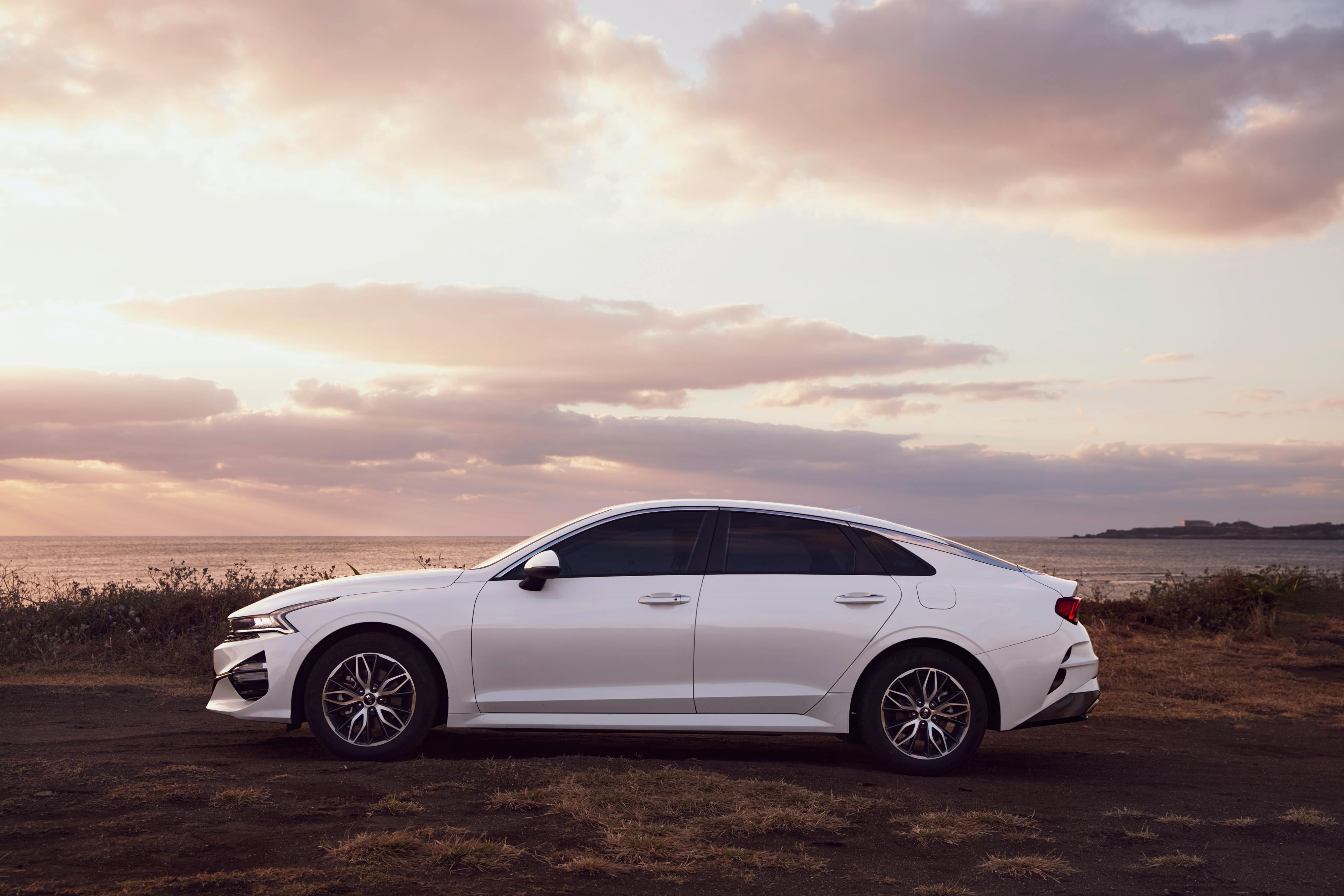 A white sedan parked on a coastline at sunset with a dramatic cloudy sky.