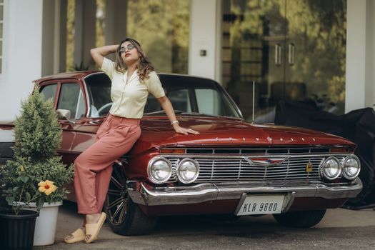 Stylish woman in vintage fashion leans against a classic red car in an outdoor setting.