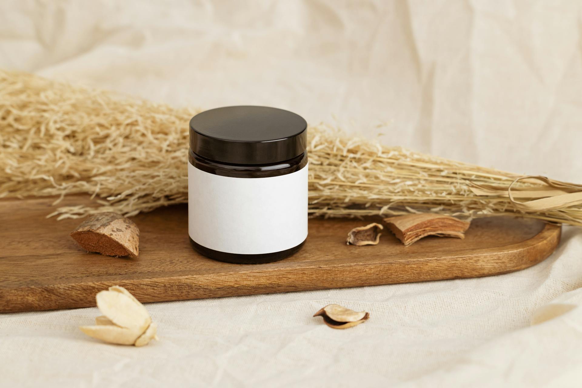 Natural cosmetic jar with blank label on a rustic wooden tray surrounded by dried herbs.