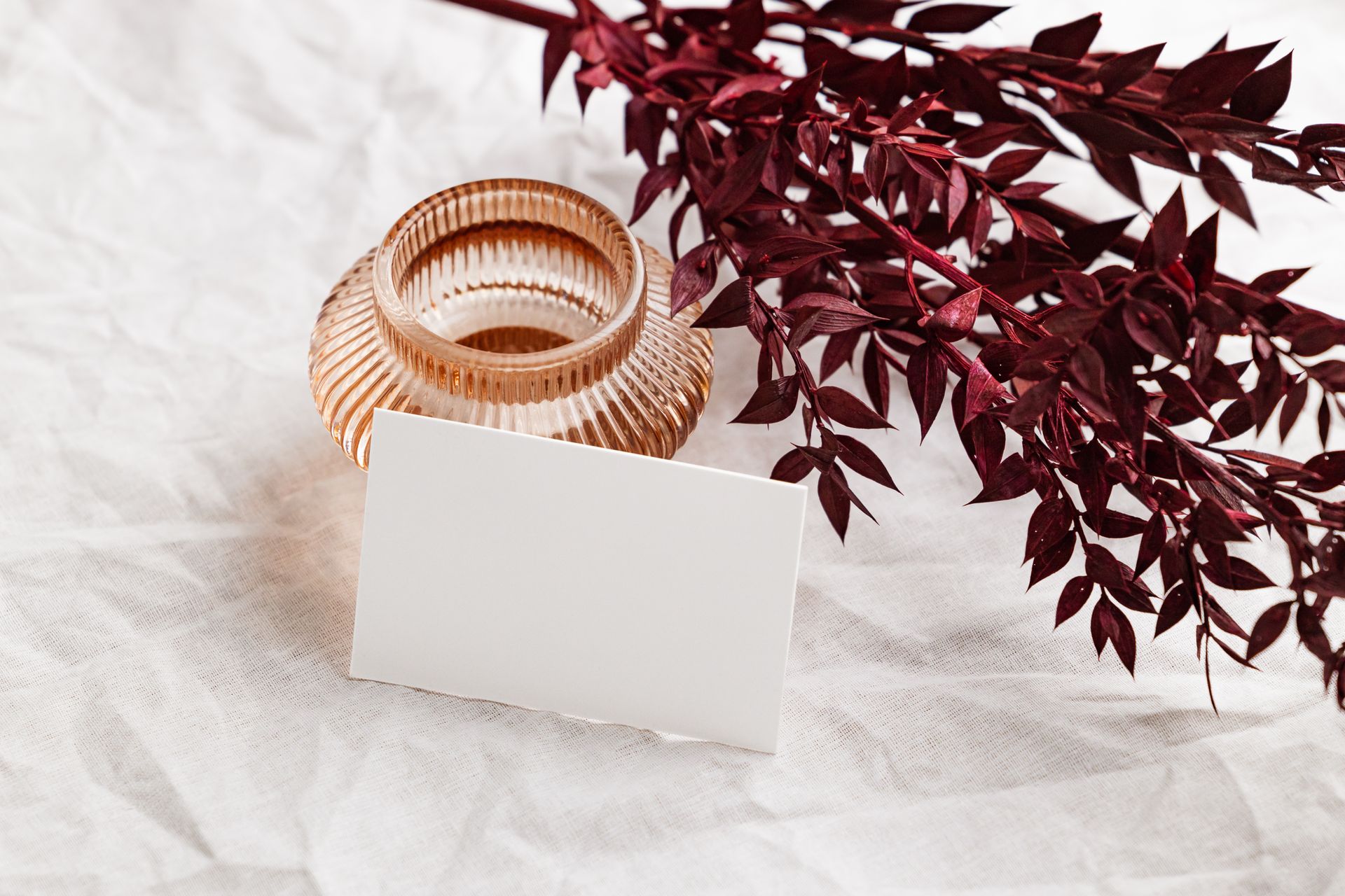 A flat lay of a blank card on textured fabric with a decorative vase and burgundy leaves.