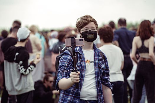Young individual wearing a mask records a crowd with a smartphone mounted on a selfie stick.