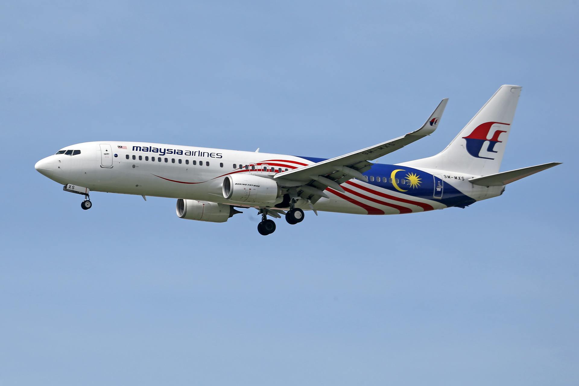 A Malaysia Airlines plane captured mid-flight, showcasing its distinctive livery against a bright blue sky.