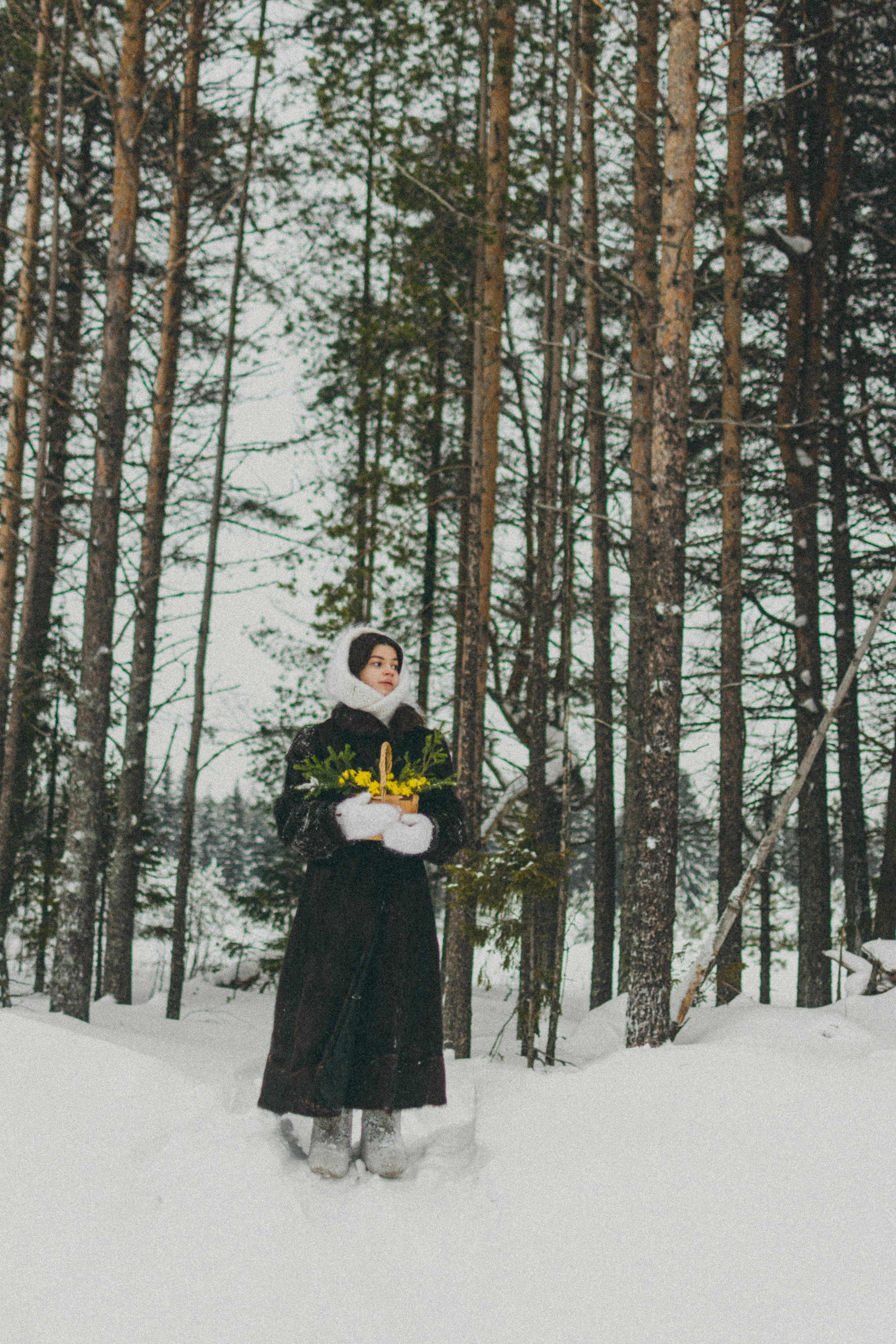 A woman in warm clothing stands in a snowy forest holding yellow flowers.