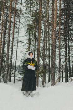A woman in warm clothing stands in a snowy forest holding yellow flowers.