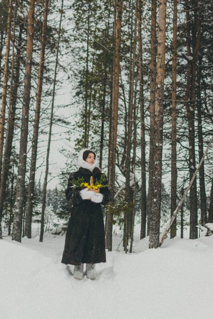 A woman in warm clothing stands in a snowy forest holding yellow flowers.