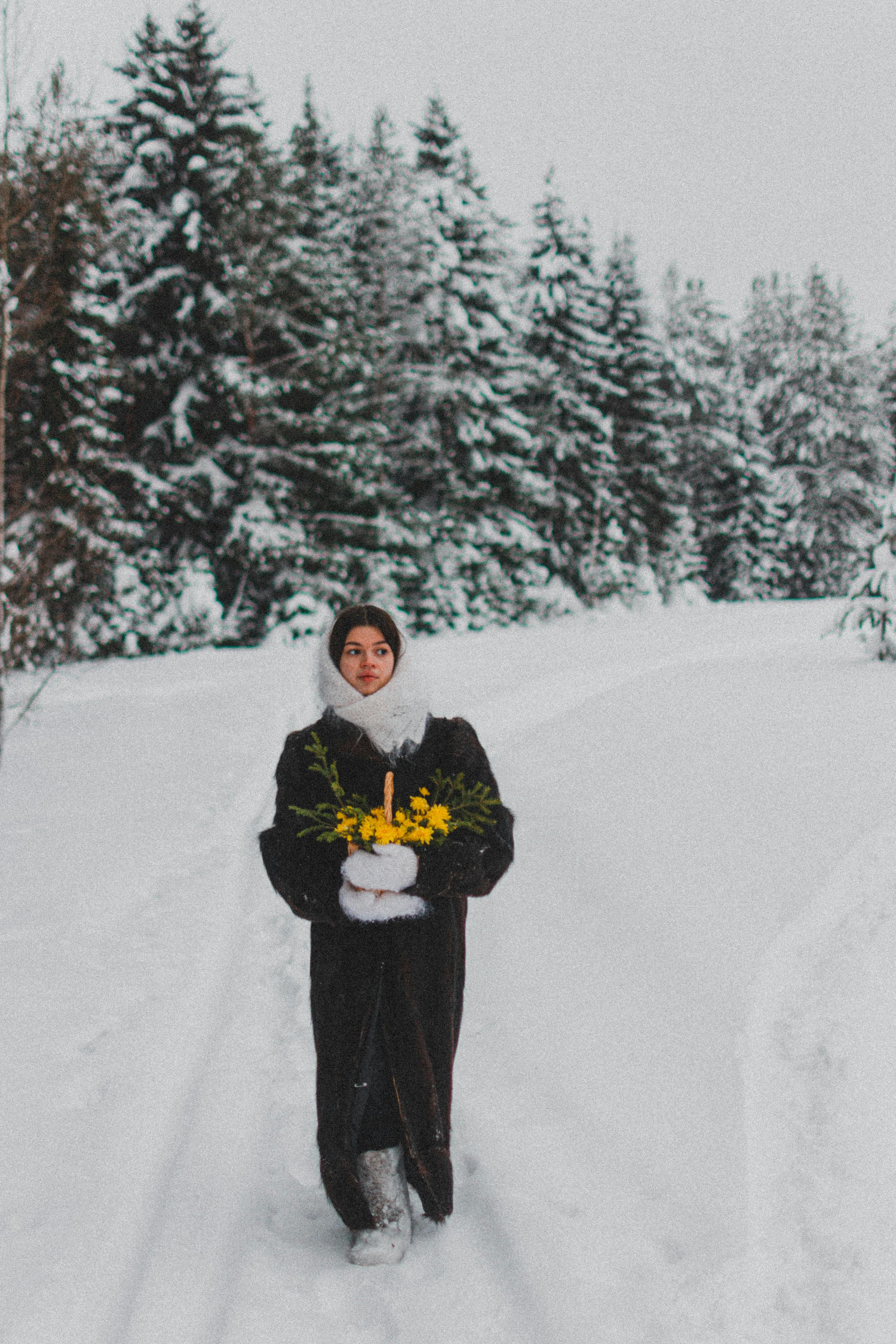 A woman in a fur coat walks through a snowy forest holding yellow flowers, evoking a winter wonderland charm.