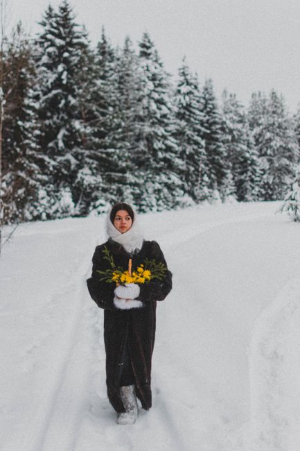 A woman in a fur coat walks through a snowy forest holding yellow flowers, evoking a winter wonderland charm.