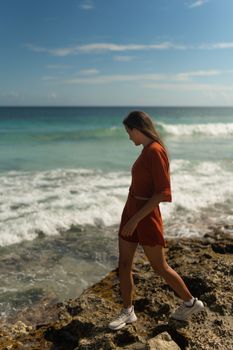 A woman in a red dress walks along a rocky beach under a clear summer sky.