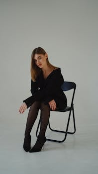 Stylish woman in a black outfit sitting gracefully on a chair in a studio environment.