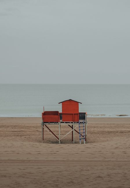 A solitary red lifeguard hut stands on a quiet, sandy beach under an overcast sky.