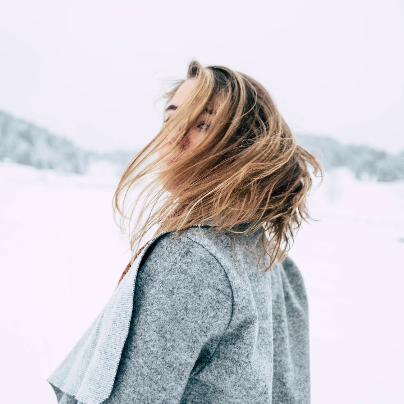 A young woman with blonde hair poses outdoors in a snowy winter setting, her hair caught in motion.