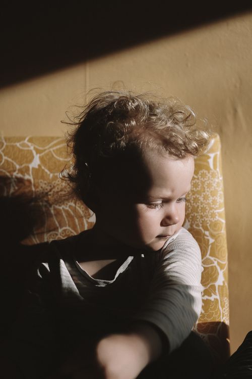 Free A young child sits in a sunlit room casting gentle shadows, creating a serene and introspective scene. Stock Photo