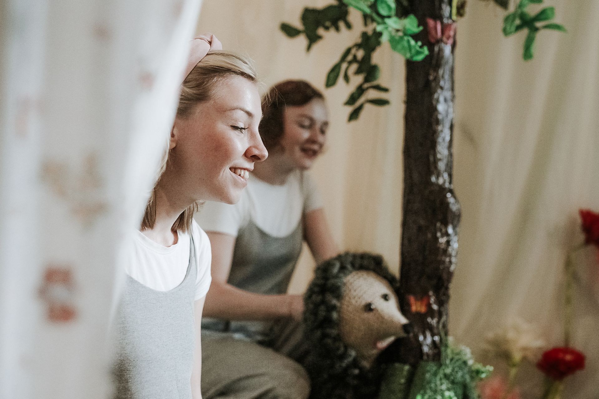 Smiling women enjoying a playful performance with handmade props indoors, featuring a hedgehog and tree.