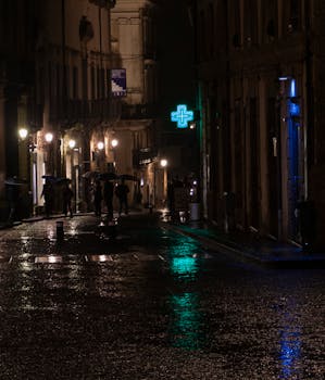 A rainy urban street scene at night, featuring silhouetted pedestrians and vibrant reflections on the wet pavement.